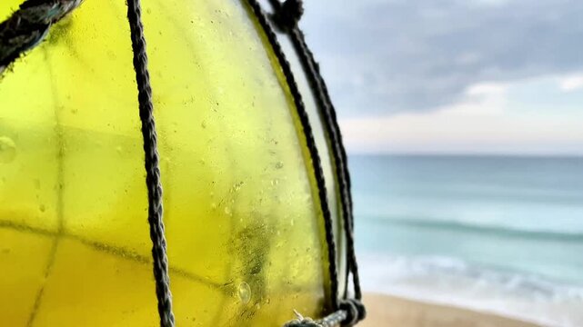  Close-up of a yellow glass fishing float in a rope net on a beach.