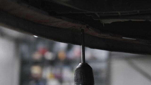 Close Up of Using a Cordless Electric Drill to Fix a Car Bumper Underbody Panel in an Auto Repair Workshop