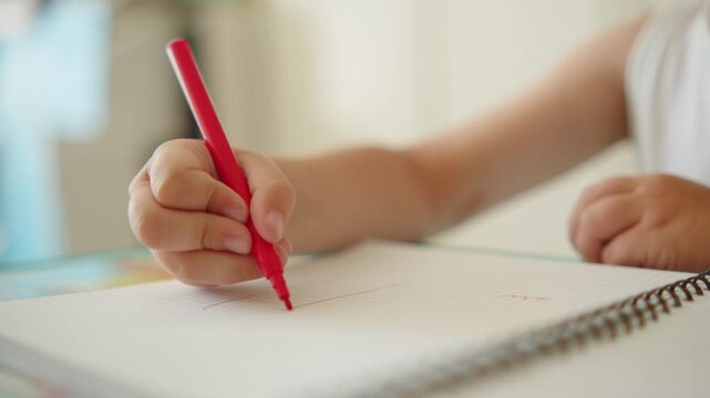 Close-up of a child's hand holding a red pencil and drawing on a blank notebook page. Little boy learning how to draw in a kindergarten. 