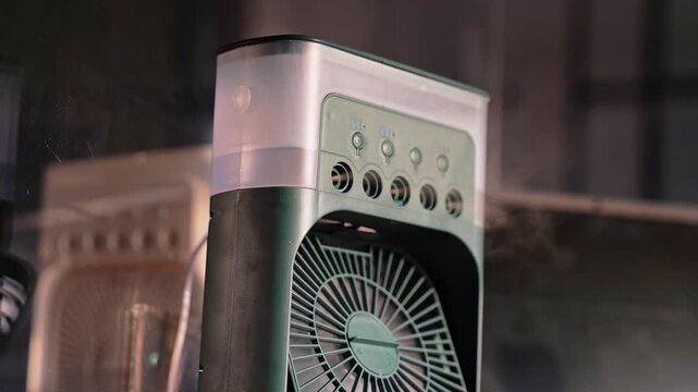 Close up of a portable desktop misting fan in a dim indoor setting. Visible vapor mist spray from a sleek air cooler device with electronic control buttons for personal cooling.
