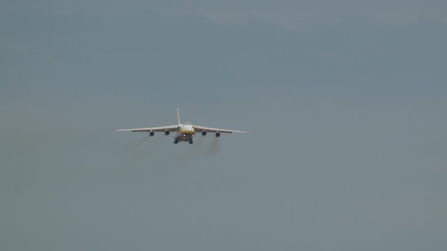 Large four-engine heavy transport aircraft approaching runway with landing gear extended, seen from the front against overcast sky