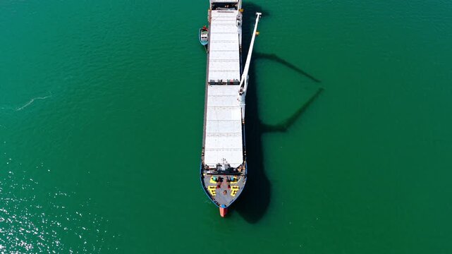 Top Down Bulk Carrier Ship Shadow on Emerald Water. A top-down drone view shows a large bulk carrier with a long crane shadow on emerald green water.