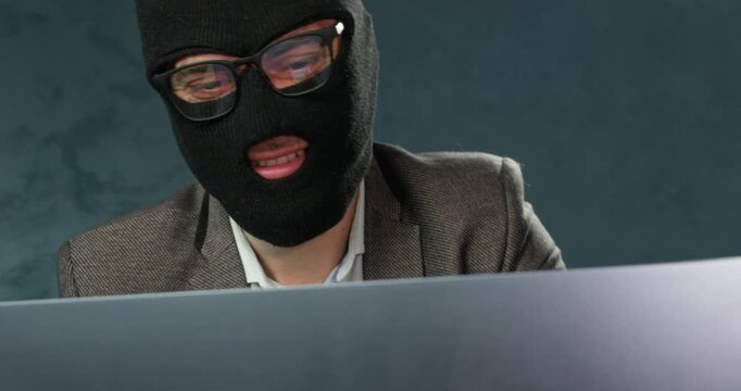 Masked scammer smiles as shows bank card while using laptop for fraud. Man in balaclava nods head presenting payment card during successful online deception activity