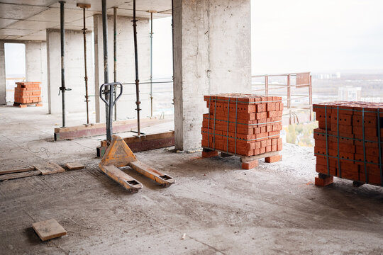 A yellow manual pallet jack and strapped stacks of red clay bricks on an unfinished concrete floor with metal shoring props at a high-rise construction site.