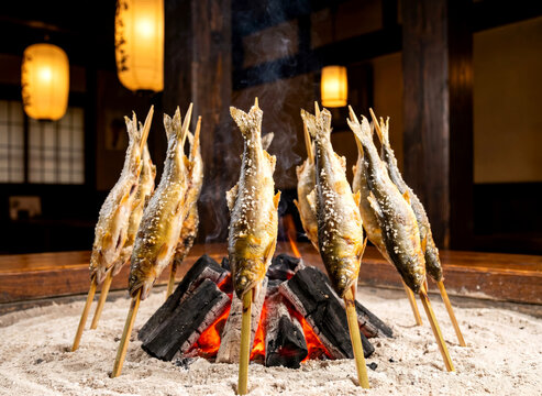 Traditional japanese ayu sweetfish on bamboo skewers grilling around a hot charcoal fire in an irori hearth with smoke.