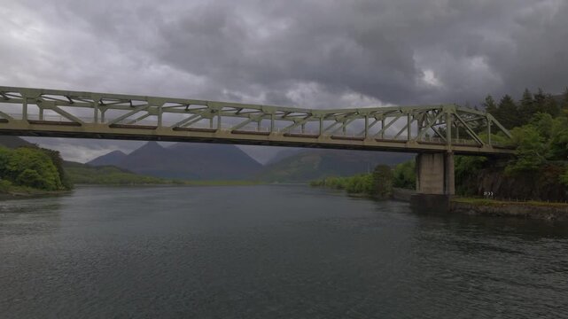 Drone backward and upward reveal from the Ballachulish Bridge, showcasing the steel span crossing the water with the moody mountains of Glencoe under stormy spring skies.