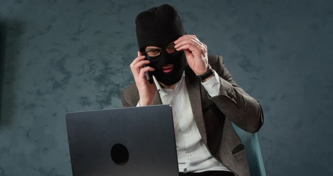 Masked scammer adjusts glasses while sitting at laptop before phone fraud. Man fixes eyewear while preparing to continue deceptive online activity in room