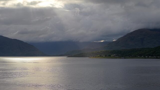 Dramatic drone view over Loch Linnhe in the afternoon, showing sunbeams bursting through heavy storm clouds to illuminate the Scottish Highland shoreline