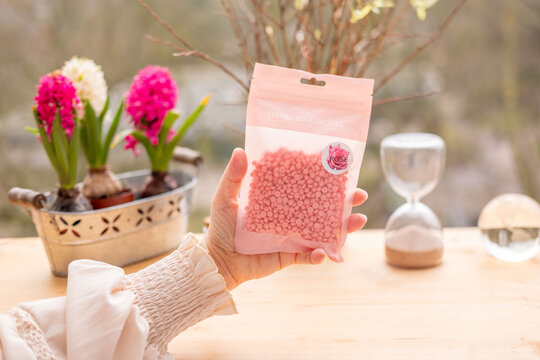Woman hand holding pink hard wax beans package for hair removal, rose scent, beauty treatment, self care, spring decor, natural feminine cosmetics, spring willow branches on wooden table, smooth skin