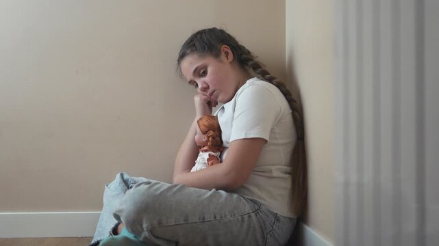 Young girl sitting on the floor with her doll. Girl punished with a doll in a corner. A teenage girl is traumatized by violence and psychological trauma. A young girl is on the floor lifestyle with.