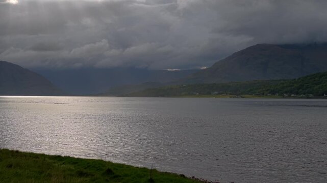 Drone flies back over grass shore as light shafts illuminate shore of Loch Linnhe on a spring evening with stormy clouds in sky