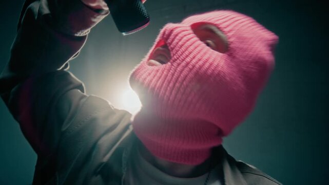 Close up shot of male hip hop artist wearing pink balaclava mask singing to microphone while recording music video backlit in studio