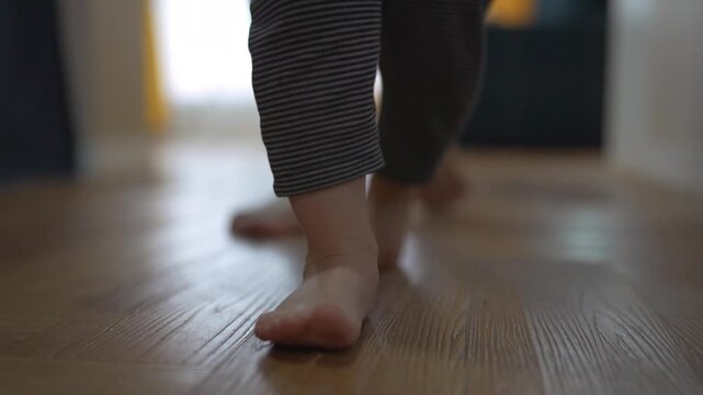 Person walking on a wooden floor. A father teaches his baby first steps. Family takes a child step in the living room. An individual lifestyle strolling on a wooden surface.