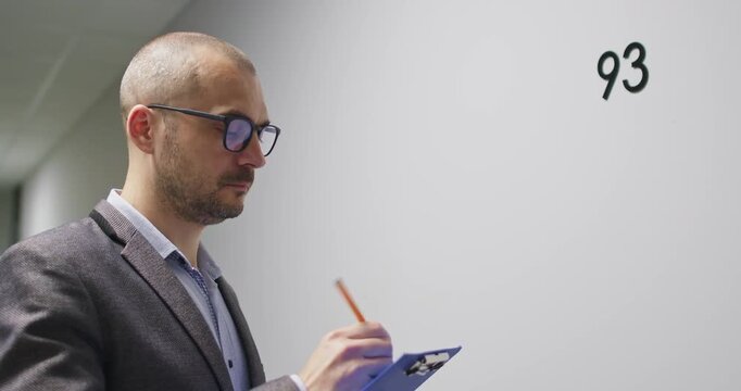 Man in glasses carefully inspects newly built apartments in modern residential building. Bald businessman writes technical details and observations into official document