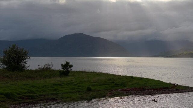 Drone flies back and up from grass outcrop as light shafts illuminate shore of Loch Linnhe on a spring evening with stormy clouds in sky