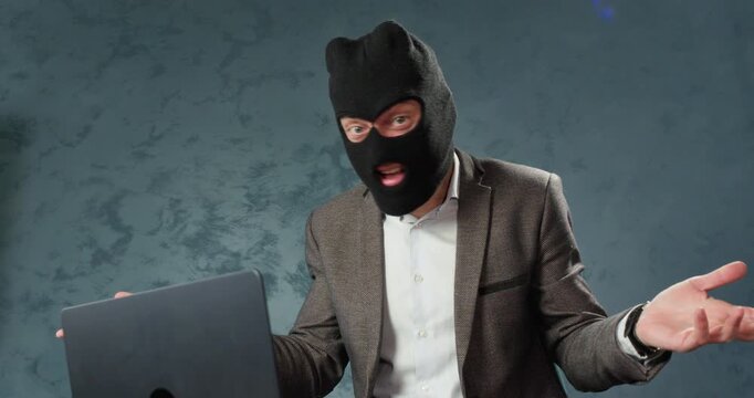 Male scammer in balaclava grabs head in frustration while sitting at laptop. Man reacts dramatically to failed fraud result while shows angry disappointment