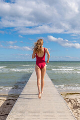Obraz premium Blond woman wearing red swim suit running on a jetty into the Baltic Sea. Creation supported by AI.
