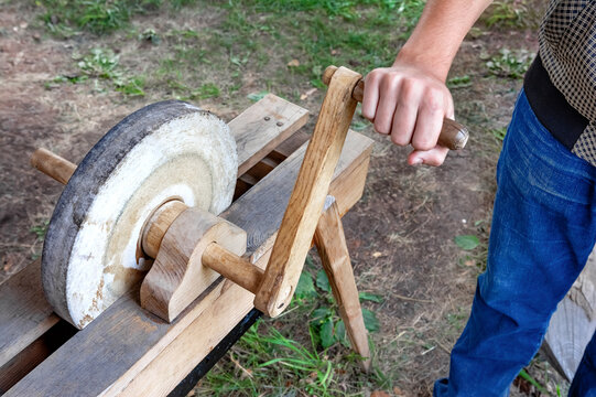 A man is whetting a tool on an antique hand-operated grindstone.