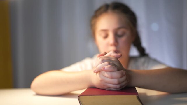 Young girl sitting at table with her hands on her head. Religious girl praying to a child. Religious book of prayer with hands lifestyle on a table. A young girl is at a table with her hands on her.