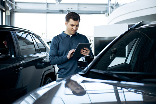 Car salesperson working in a dealership showroom, standing between vehicles and using a digital tablet for inventory management