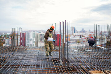 A construction worker in a hard hat carrying a coil of wire walks across a vast steel rebar grid preparing for concrete pouring on a high-rise building site with a city view. © Aigul Sabirova