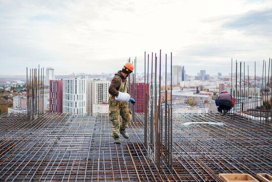 A construction worker in a hard hat carrying a coil of wire walks across a vast steel rebar grid preparing for concrete pouring on a high-rise building site with a city view.