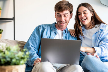 Young couple sitting on a sofa at home smiling and looking at a laptop together, enjoying online...