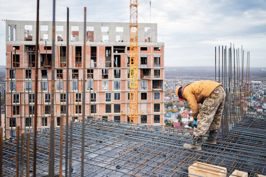An ironworker bends to tie a steel rebar grid on a high-rise concrete slab, with a brick building under construction and a yellow tower crane in the background.
