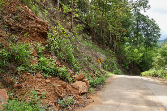 Steep landslide slope natural disaster erosion danger on rural concrete road edge surrounded by green forest trees under cloudy sky creating desolate and alarming environmental hazard
