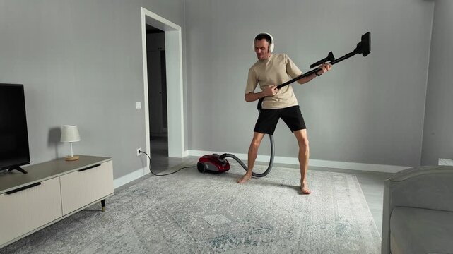 Energetic barefoot man having fun cleaning the house. He is listening to music on headphones and pretending to play a guitar with a vacuum cleaner