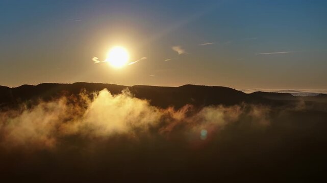 Drone aerial shot of sunrise above mountain ridge, dense illuminated fog glowing in warm golden light in foreground, blue sky above, lens flare