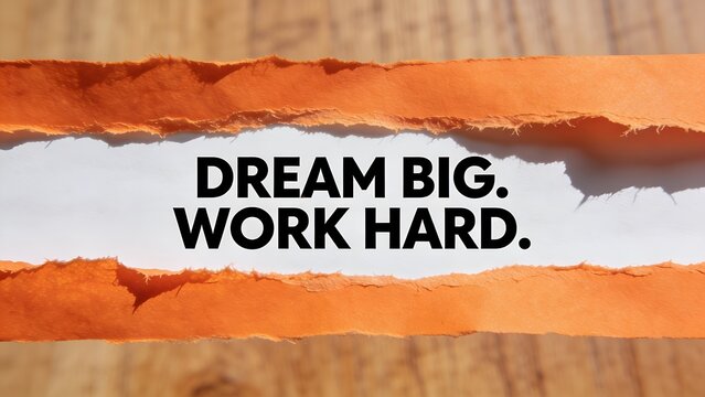 Bold black motivational text reading dream big work hard printed on white paper revealed behind torn edges of vibrant orange construction paper in bright natural lighting