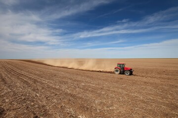Naklejka premium The Tractor Tilling Vast Dry Farmland Under Dramatic Blue Sky With Dust Trail