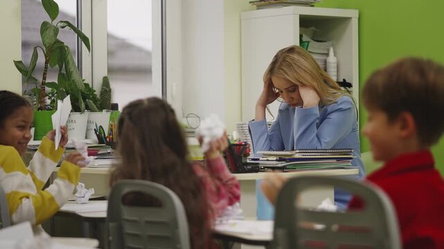 Exhausted teacher holding head while pupils throw paper planes during lesson