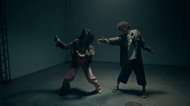 Theatrical dance performance shot of two young dancers passing wireless speaker while doing emotional hip hop freestyle choreography in empty industrial studio lit by flashing lights