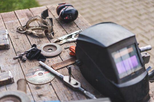 Assorted workshop tools including welding helmet and cutting discs placed on a wooden stool outdoors. Industrial DIY and metalworking equipment in natural light.