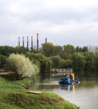 Technical industrial dredging boat cleaning a river water near green trees with factory chimneys in the background