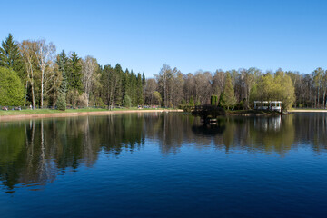 Fototapeta premium Tranquil park pond with a white gazebo on the shore during a bright sunny day. Beautiful landscape photography of a natural water reservoir suitable for travel and tourism banner.