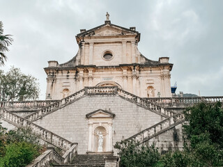 Fototapeta premium Grand Stone Steps Leading Kotor Church, Baroque Facade Framed By Overcast Sky, Worn Marble Balustrades, Empty Plaza With Cobblestone Pavement,