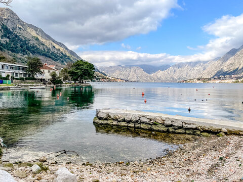 Pebbled shore with distant mountain panorama, shallow clear water lapping rocky edge, moored buoys marking calm swimming area,