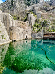 Ancient Kotor Fortress Beside Emerald Water. Massive Stone Ramparts Curve Above Calm Moat With Turquoise Reflections, Wooden Covered Quay On Right, © Наталья Северова