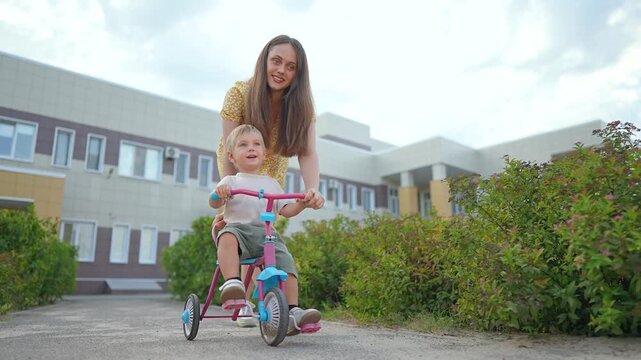 Mother helps little son ride tricycle on street. Happy boy holds handlebars while mom pushes bike. Parent teaching kid to ride outdoors. Mother and son enjoy active family time together on walk