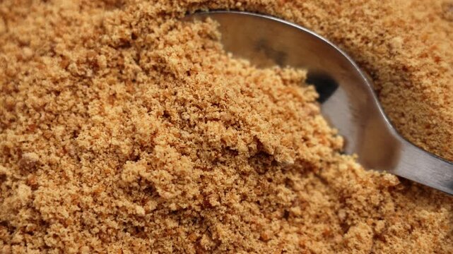 Brown cane sugar with stainless steel spoon close up. Unrefined grainy crumbly sweet substance. Top view. Rotation