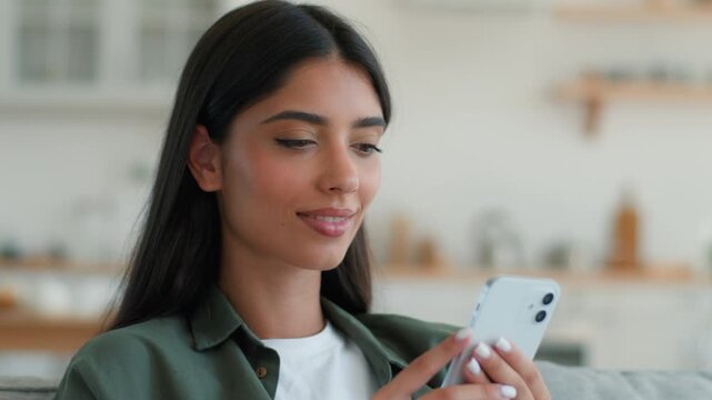 Pensive smiling Arabian Indian woman using smartphone at home indoors thinking dreaming purchase shopping online chatting social media app relaxed happy female holding mobile phone pondering decision