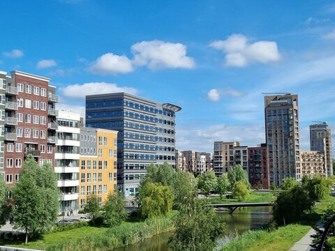 Scenic urban landscape in Zuidas, South Amsterdam, featuring colorful residential and office buildings along a sustainable water canal with green banks under a blue cloudy sky. No people, copy space.
