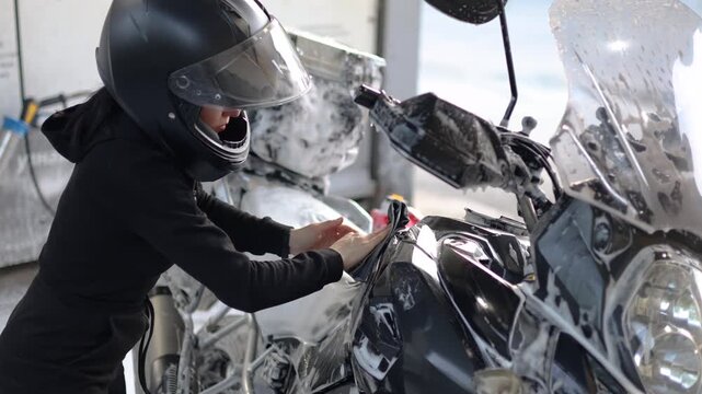 Close-up slow motion 4k footage of a woman in a black jumpsuit and helmet washes her motorcycle with a rag after applying foam at a self-service car wash. Woman takes care of her motorcycle.