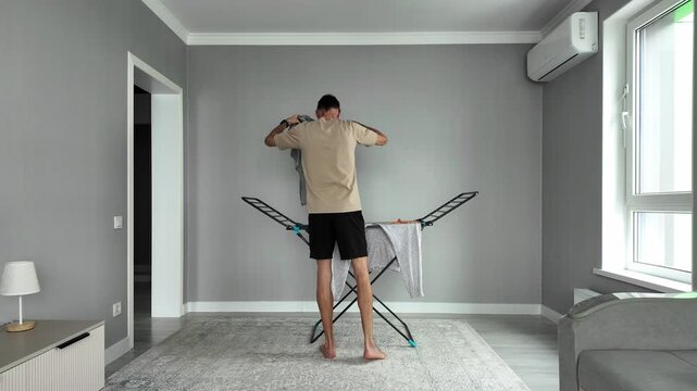 Millennial male doing laundry household chores in a modern living room. Housekeeping routine of a single man hanging clean clothes to dry