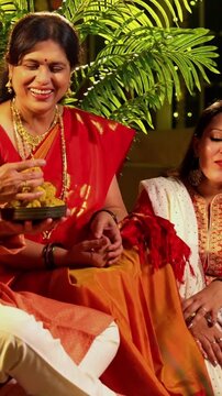 Holding laddus and smiling together, Indian family with young girl, senior mother in saree, father and husband in traditional wear celebrates Diwali indoors with diya, flower rangoli, and laughter
