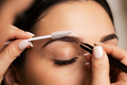 Close-up of a beauty therapist using tweezers and a spoolie brush to shape and style a woman's eyebrows during a professional brow lamination or grooming session