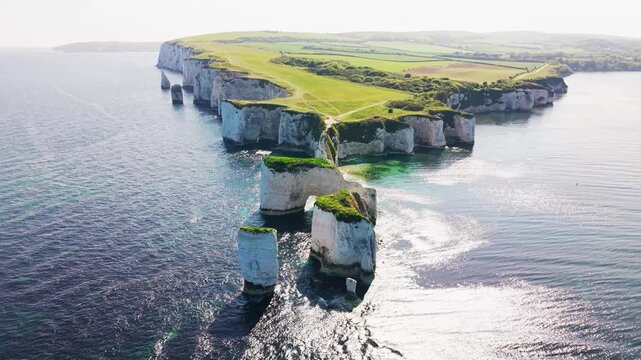 Aerial drone footage of Old Harry Rocks on the Dorset coastline in England, showing dramatic white chalk sea stacks, cliffs, turquoise water, and green clifftop paths. The shot captures the famous